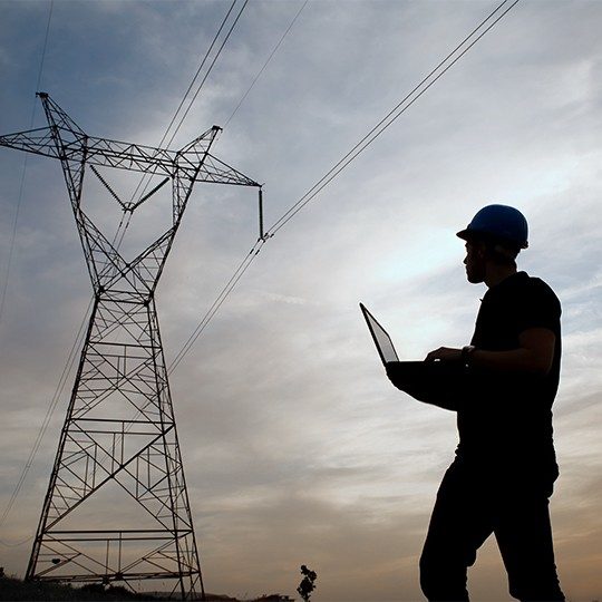 Network engineer working in the field, wearing a hard hat and holding a laptop, looking at a network tower at dusk 